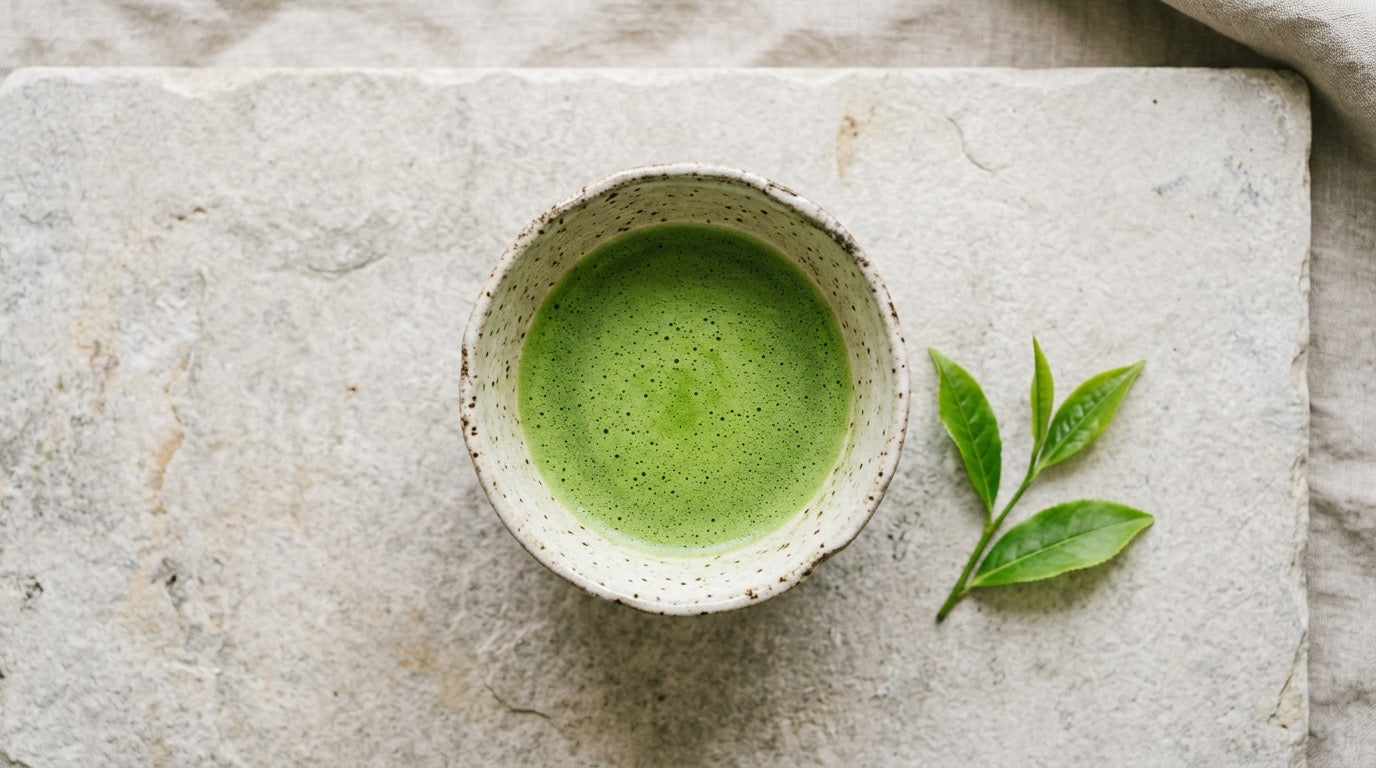 A bowl of bright green ceremonial matcha with fresh tea leaves on pale stone, a calm healthy scene.